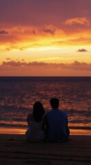 Fototapeta premium Couple sits on beach watching vibrant sunset over ocean. Waves gently lap shore as sky glows with orange and yellow hues. Peaceful moment shared by two people.