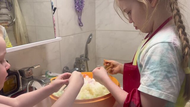 A child is mixing a salad in the kitchen sink. Another child is helping with the ingredients. Both are wearing bright aprons. The scene demonstrates teamwork and fun. Perfect for family cooking or cul