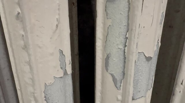 Peeling paint reveals aged, worn white radiator panels. Faint cracks show signs of time and weather exposure. Dark gaps between pipes hint at old, unused heating system