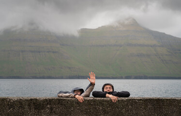 Fototapeta premium Humorous concept of a young couple in raincoats pretending to sink and reaching for help against misty mountains in the Faroe Islands