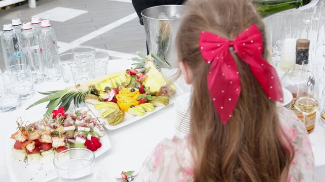 Little girl with beautiful red hair accessory looking at a catering buffet table with fruit platter