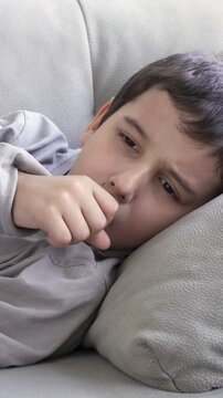 Sick child feeling unwell at home, coughing into fist while lying on couch under white blanket with tissues nearby, ill male kid resting during the day in quiet living room