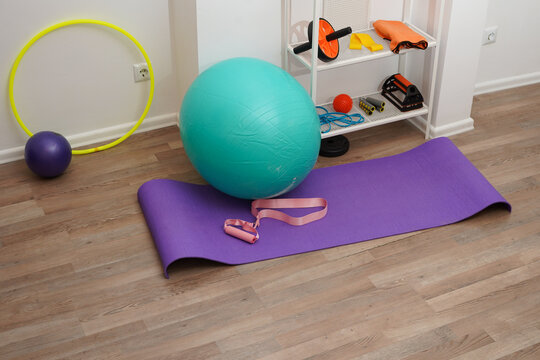 A collection of colorful exercise equipment for a home workout or physical therapy. A purple yoga mat, turquoise stability ball, and resistance bands are on a wooden floor in a bright, modern room.