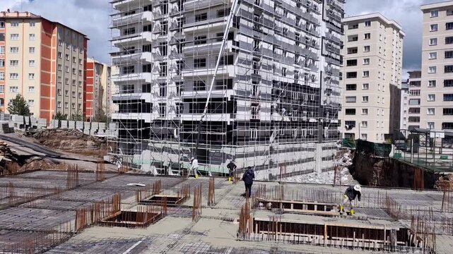 Wide horizontal static view of a residential construction site layout featuring an extensive steel rebar foundation grid and a scaffolded high-rise building.