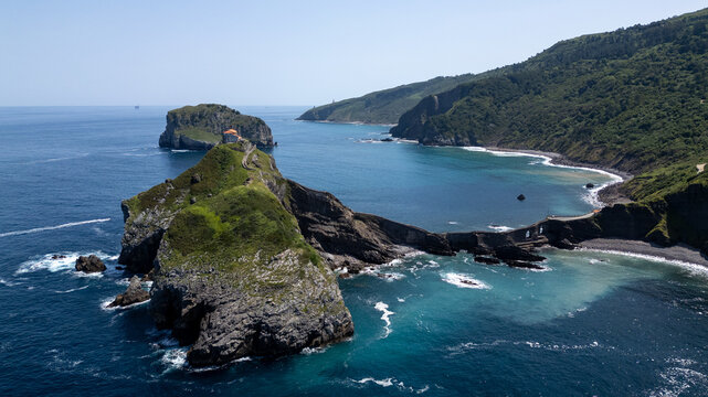 Aerial drone view of Gaztelugatxe, a small rocky island connected to the mainland by a historic stone bridge and staircase. Blue waters of the Bay of Biscay. Impressive cliffs, rugged landscapes, and 