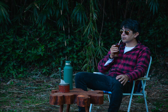 joven con lentes de sol y camisa a cuadros roja y negra bebiendo un mate caliente en el parque, expresi&oacute;n de tranquilidad y calma