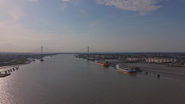 Dartford Crossing and oil tanker ships on river Thames aerial view. Afternoon sun reflection on water surface. Cables stayed bridge with moving traffic. Oil terminal with storage tanks.