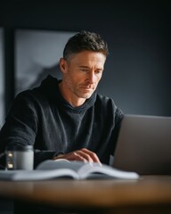 Obraz premium Focused man working on laptop with book and coffee in modern office