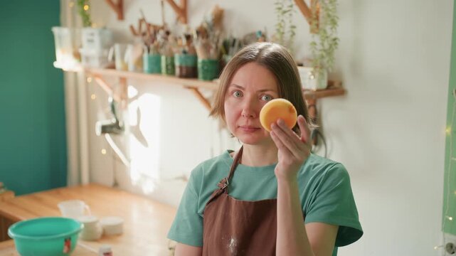 Caucasian potter in workshop holding two sponges and small bowl, playful demonstration of texture and absorbency, bright green bowl and shelves of glazes visible, expressive instructional moment