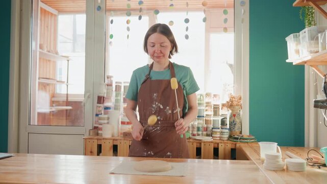 Calm white woman potter prepping clay on wooden table, wearing paintstained brown apron, sunlight through window, shelves of glaze jars, focused hands sponging surface, gentle water droplets, soft