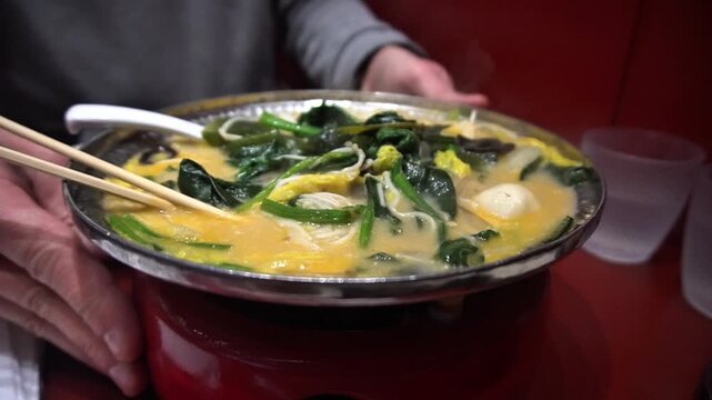 Man turns a hot pot of Chinese soup with spinach, seaweed and noodles. Delicious hot Asian soup with vegetables and seafood for lunch close up
