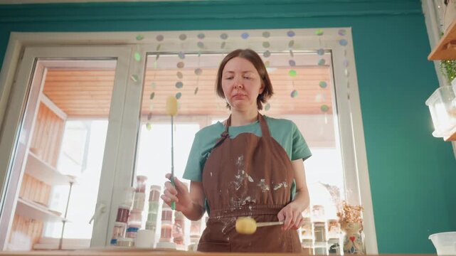 Pottery workshop artist prepping glaze in bright studio with jars on shelves, apron splattered, applying sponge strokes to bisque pieces, calm focused morning mood, sunlit window backdrop