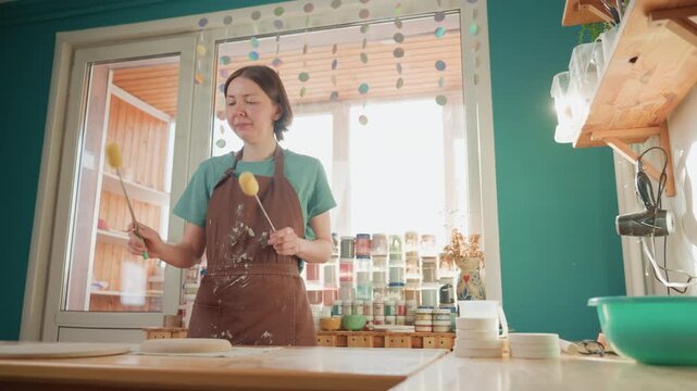 Pottery workshop maker playfully sponging glaze on wooden table surrounded by stacked jars, colorful splatter and lively motion, hands tapping brush, green bowl and sink visible, playful experimental