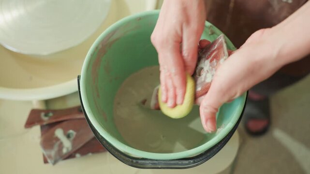 Hands cleaning pottery tool in bucket, closeup of sponge and metal rib by wheel. Quiet studio scene showing artisan rinsing clay residue, gentle circular motions, green basin and wet hands, mindful