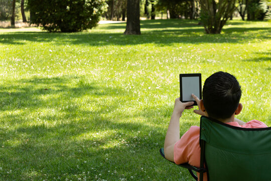 A young man turns the page of a touchscreen e-book while sitting in a chair in a park on a sunny day