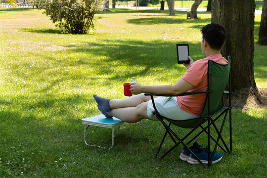 A young person is relaxing in the park with an e-reader and a drink