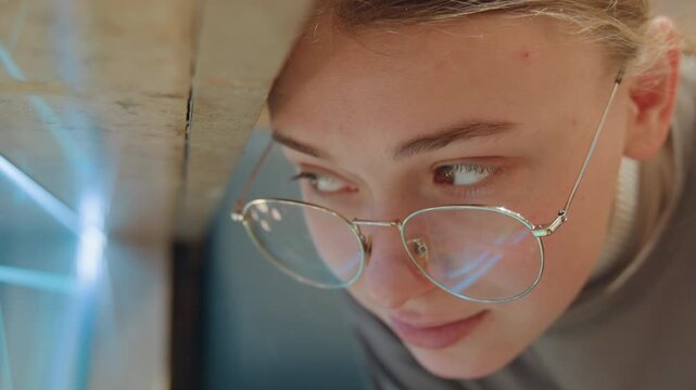 Young caucasian woman peering through glass, closeup portrait with round glasses and chain, soft neon reflection on lens, freckle and subtle smile, indoor evening mood, art student sketching,