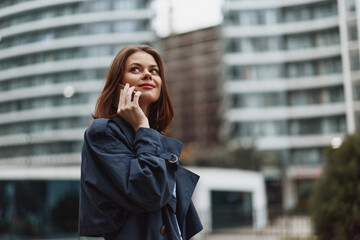 Fototapeta premium A thoughtful woman in a coat stands outdoors in an urban cityscape, talking on a phone, with modern buildings behind, capturing casual communication in daily life.