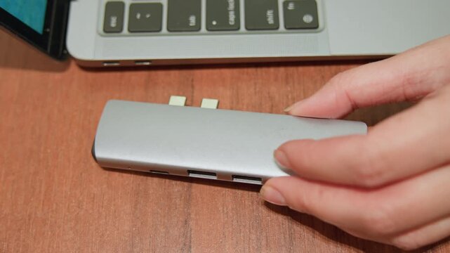 Silver hub beside laptop keyboard, hands attaching aluminum multiport adapter to laptop edge on wooden desk, designer setting up compact docking station for productivity, closeup steady motion,