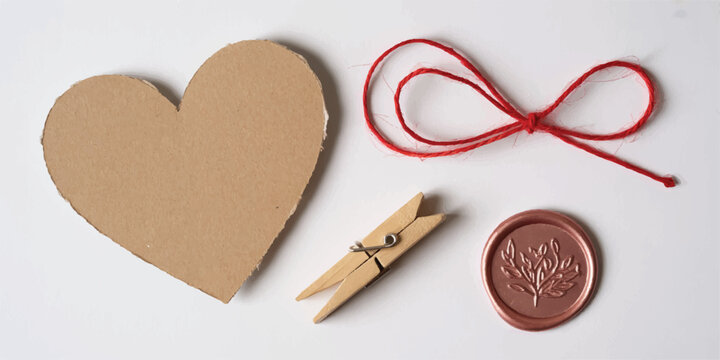 Rustic craft composition featuring cardboard heart red string bow wooden clothespin and rose gold wax seal on white background.