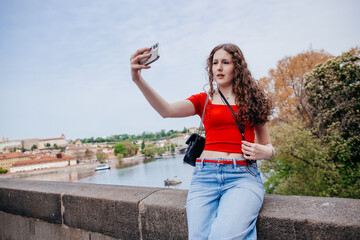 Young woman taking selfie with smartphone while sitting on bridge overlooking river city