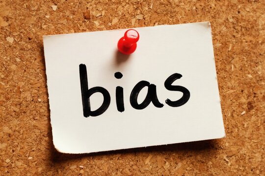 White paper note with word bias handwritten in black ink pinned to corkboard. Word represents prejudice, unfair opinion, or leaning towards one side, needs careful consideration.