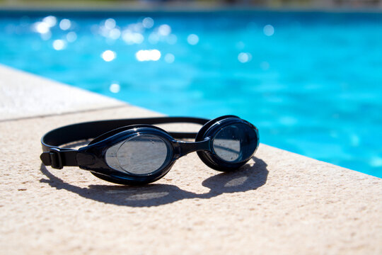 Black swimming goggles resting on poolside with blue water background