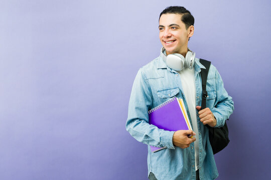Smiling male university student looking away, holding notebooks and wearing headphones, backpack, education concept