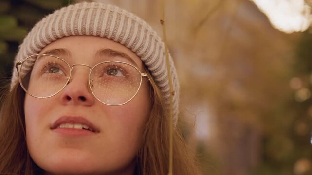 Young white woman in beanie wandering among pine branches under warm sunset, glasses catching bokeh light, contemplative botanist vibe with gentle sparkle on lenses and cozy golden tones
