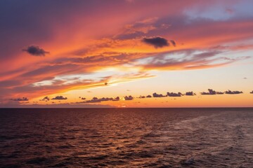 Obraz premium Dramatic sunset over calm Baltic sea water. Orange pink clouds streak across vast sky, sun peeks at horizon. Ripples on ocean surface reflect twilight colors. Serene evening seascape.