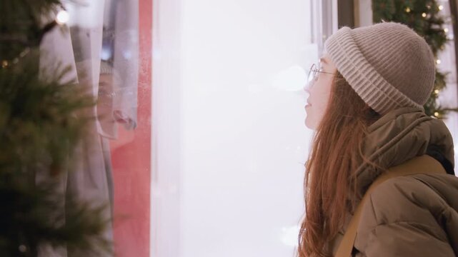 White woman window shopping by display, reflective glass reveals mannequin silhouette, contemplative gaze in holiday evening, beanie and puffer coat, urban storefront glow, nostalgic mood