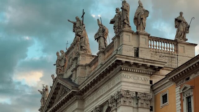 A wide shot of the grand Saint John Lateran Basilica in Rome, Italy, showing its beautiful Baroque facade and statues under dramatic clouds with tourists in the foreground.

