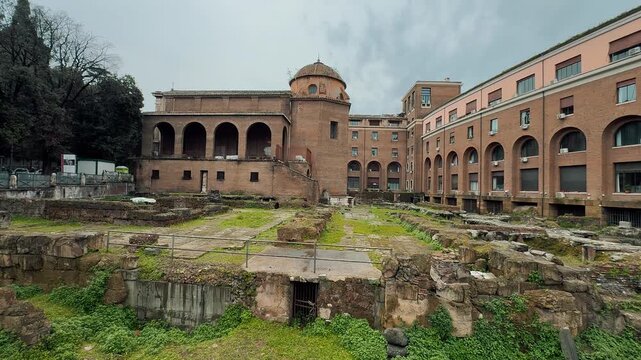 A scenic wide shot of the ancient Roman ruins and the historic brick Baptistery of Saint John in Lateran under a dark, moody, cloudy sky in Rome, Italy.

