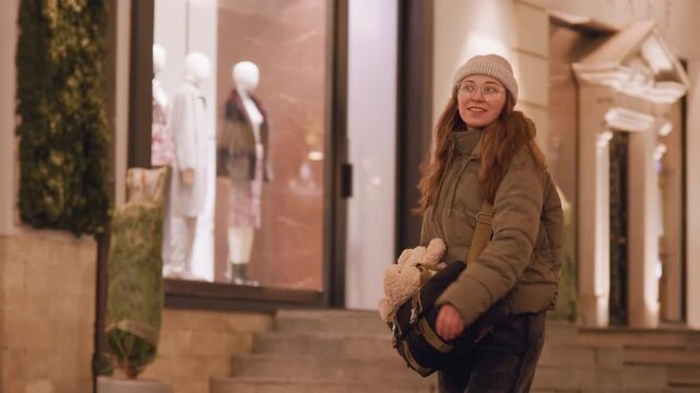 White woman outside boutique window at night, holding plush toy and gazing at mannequins on elegant steps, nostalgic shopper in puffer jacket enjoying warm holiday storefront glow and soft city