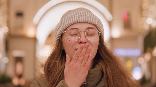 White woman smiling in festive closeup, playful friend sending kiss and making heart gesture, round glasses and knit hat, warm golden bokeh, intimate cozy portrait, cheerful expression