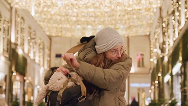 Full shot white woman holding backpack and plush toy, adjusting beanie then smiling, playful shopper vibe in holiday market, warm lights and decorated storefronts, confident stride in puffer coat