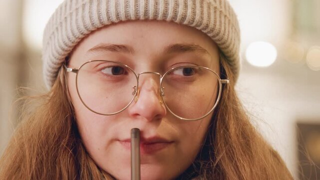 Young white woman at winter cafe sipping through straw wearing round glasses and knit beanie, closeup portrait with warm bokeh lights student sampling seasonal drink, writer pausing midnote, artist