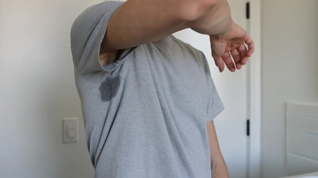 Person raising an arm showing a visible underarm sweat stain on a gray shirt, highlighting perspiration and hygiene issues in a realistic everyday context.
