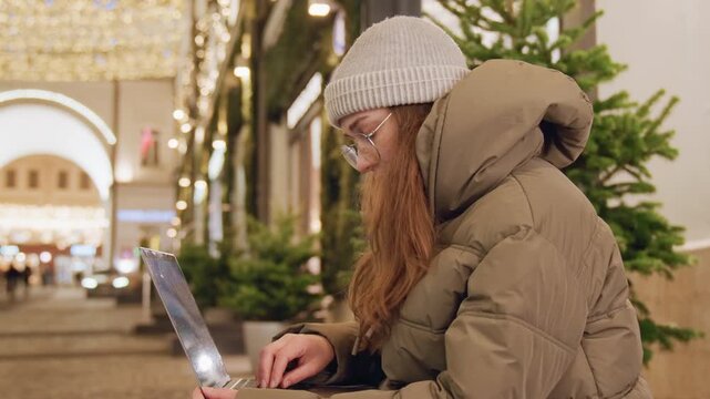 White woman freelancer working on laptop near illuminated christmas tree, bundled in beanie and puffer coat on bench, typing with focused expression against warm holiday bokeh, digital nomad vibe