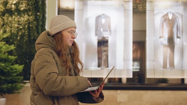 Young woman typing on laptop near storefront display, beanie and puffer jacket, evening lights and mannequins behind glass, remote work on sidewalk, intent expression and steady typing, urban holiday