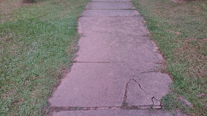 Obraz premium Worn concrete sidewalk with visible cracks and uneven joints running through grassy park surroundings