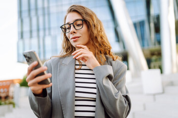 Obraz premium Portrait of a smiling woman with a phone against the backdrop of a modern city building. A stylish woman in glasses uses a smartphone outdoors. Concept of style, fashion, and business.