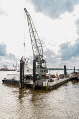 Obraz premium Industrial floating crane platform anchored on a wide harbor river with steel lifting structure, mooring posts, cloudy sky, and maritime port equipment.