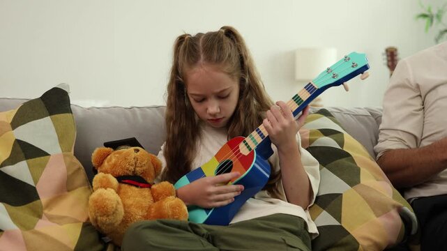 A young girl with pigtails playing a colorful ukulele on a couch, with a teddy bear and a man sitting beside her in a cozy living room.