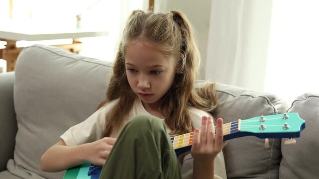 A young girl with blonde hair playing a colorful ukulele while sitting on a gray couch in a bright room.