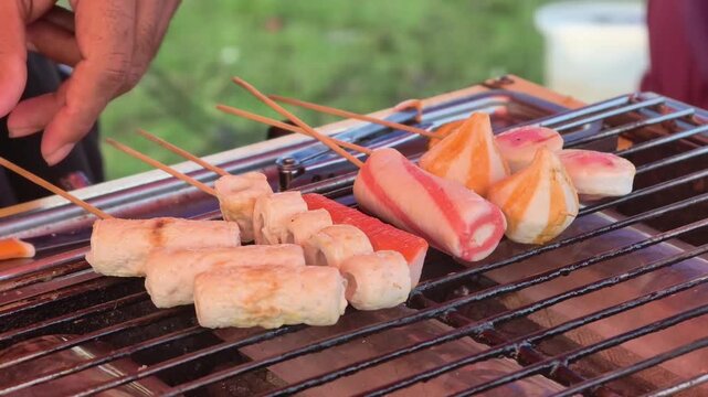 Close up of a street food vendor brushing sauce on various fish cake and sausage skewers on a hot charcoal grill.