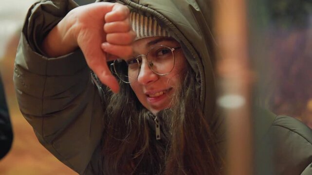 Young woman in hood shows thumbsdown at night transit shelter, beanie and glasses, dismissive gesture, neon bokeh lights, subtle frown, grainy film texture, cold jacket, candid critic vibe