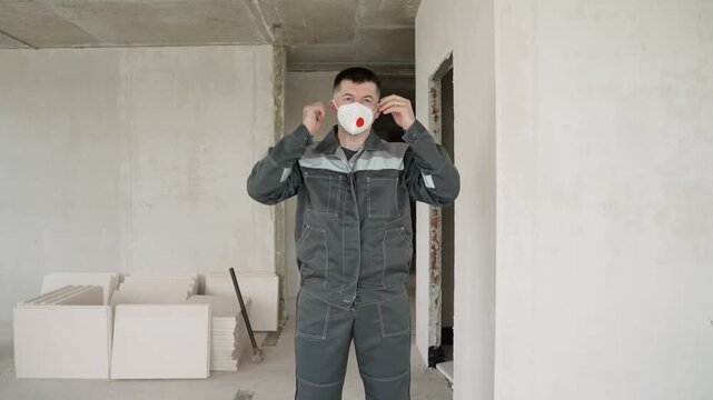 Male builder in grey uniform stands in room with white walls and tiles, holding face mask, renovation site, safety equipment, industrial setting, neutral colors
