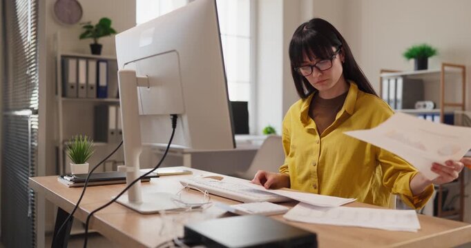 Woman freelancer reviewing documents at computer. In a tidy home office she works remote, checking notes and figures with focus at a spacious desk. Concept of focused remote productivity.
