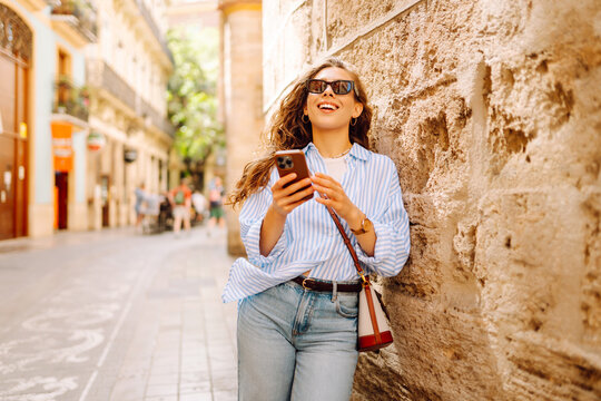 A young woman with a phone enjoys the sunny city streets. A tourist with a phone strolls and poses at sunset. Concept: style, fashion, urbanism. Lifestyle.